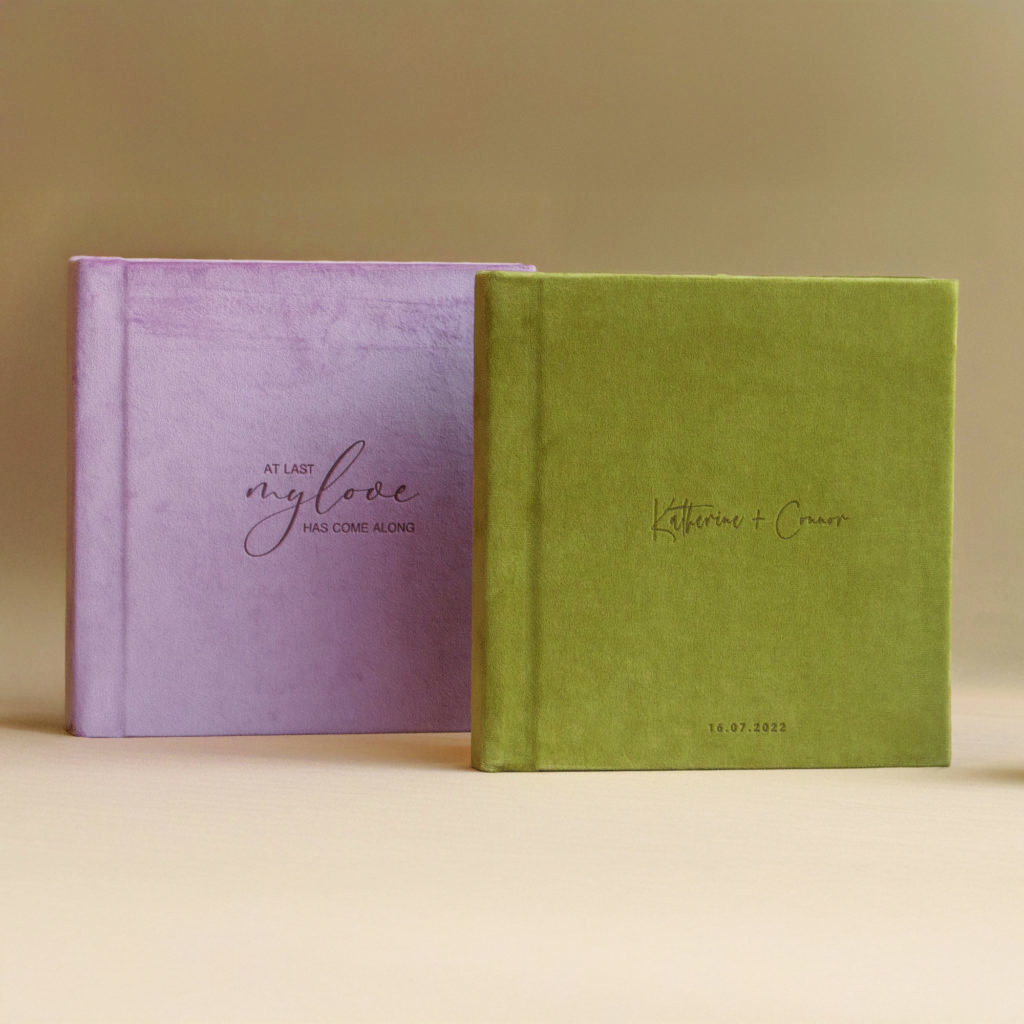 Two velvet-covered wedding photo books, one pink, one green, with personalized laser-etched covers, displayed side by side on a neutral surface.