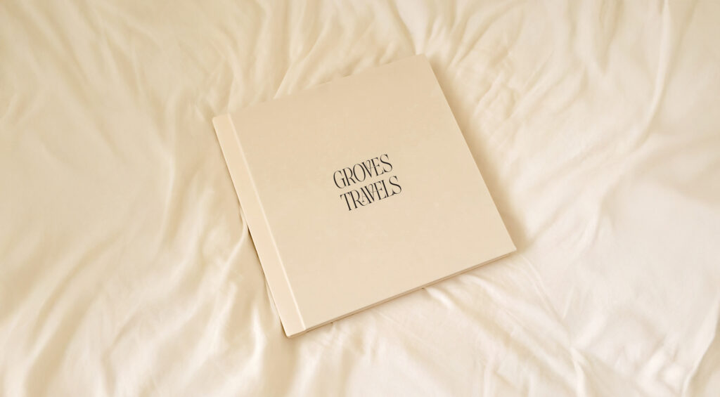 Ivory linen photo book cover with UV-printed title “Groves Travels” resting on a soft beige bedspread.