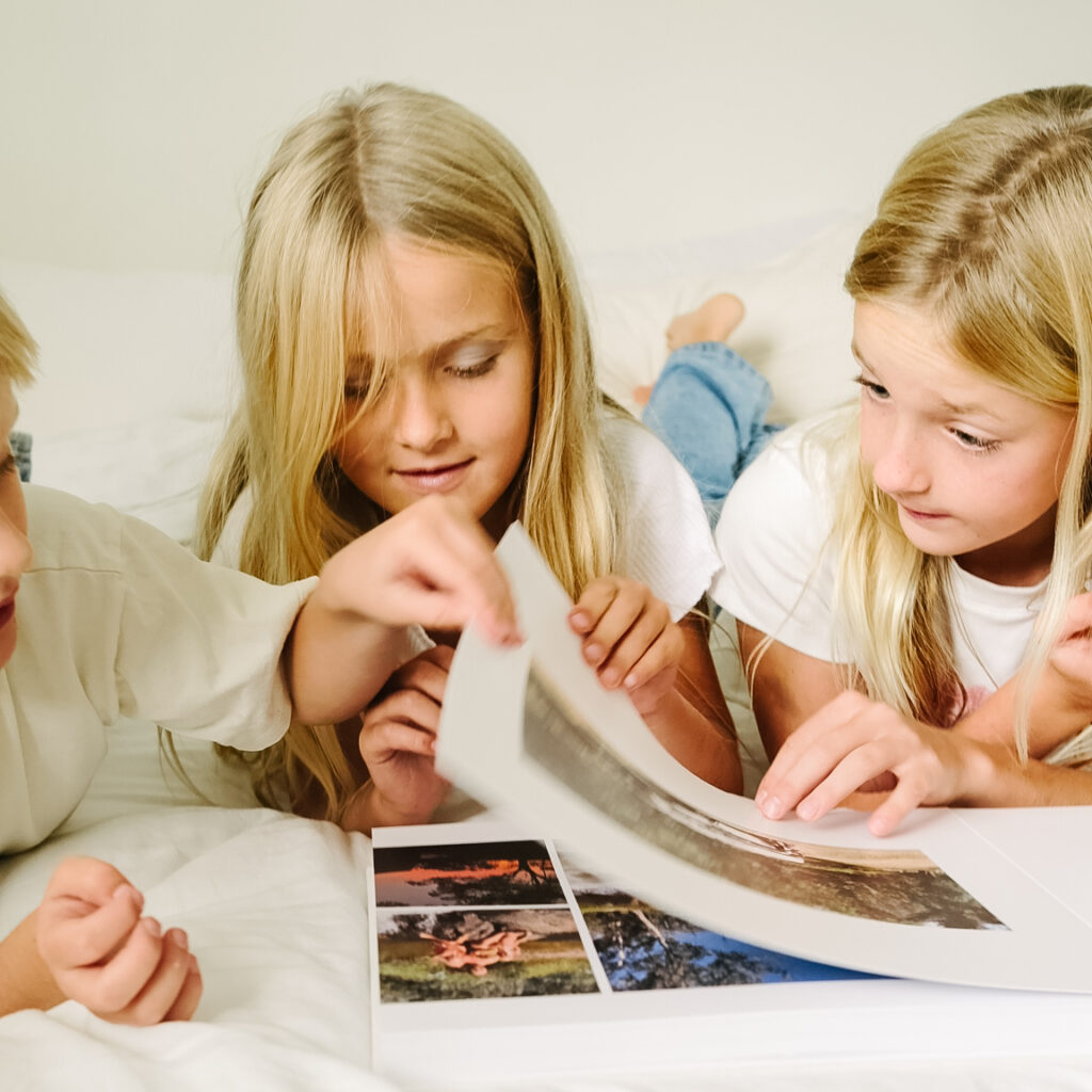 Three children flipping through a travel photo book with thick layflat pages on a white bed.