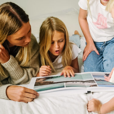 Mother and children gathered on a bed, flipping through a layflat travel photo book with beach images.