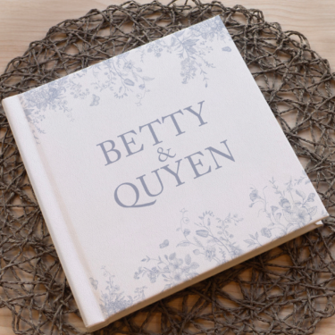 Custom white leather wedding album with UV-printed floral design and personalized title “Betty & Quyen,” displayed on a wooden table with a woven mat.